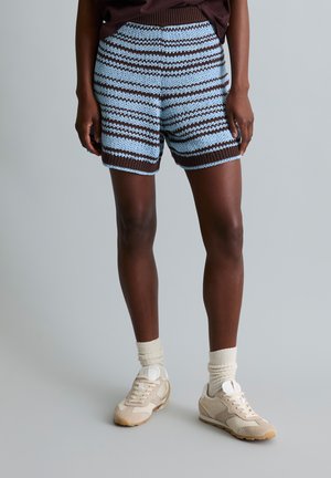 Person trägt blau-braun gestreifte Strickshorts, beige Sneakers und cremefarbene Socken vor einem einfarbigen grauen Hintergrund.