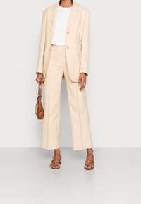 Bežový upravený blazer s klopami, spárovaný s odpovídajícími kalhotami s širokými nohavicemi. Pod ním bílý tričko, doplněné hnědou kabelkou a sandály na pásky.