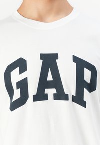 Egy fehér pólót viselő személy, amelyen nagy, vastag fekete betűkkel a "GAP" felirat áll a mellkason.