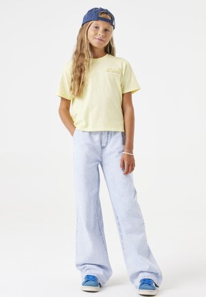 Geel cropped T-shirt met 'Love' opdruk, lichtblauwe wijde jeans en blauwe sneakers. Basebaldop in donkerblauw. Casual outfit.