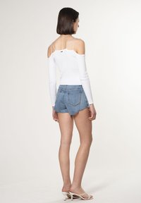 Top bianco a maniche lunghe con design a spalle scoperte, abbinato a pantaloni corto di denim blu chiaro con orli sfrangiati. Modella che indossa sandali bianchi con tacco.