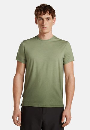 Junger Mann trägt ein schlichtes hellgrünes kurzärmeliges T-Shirt und schwarze Hose, steht vor einem weißen Hintergrund.