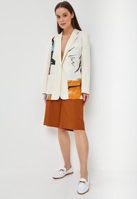 Un blazer crema con motivi astratti in blu, nero e arancione, abbinato a una gonna color ruggine e mocassini bianchi con accenti dorati.