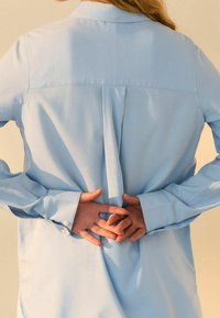 Camicia azzurro chiaro con maniche lunghe, una piega sul retro e polsini con bottoni. Il tessuto sembra liscio e leggermente testurizzato.