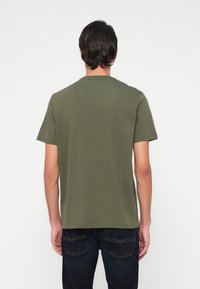 Mann med mørkt hår iført en olivengrønn kortermet T-skjorte og mørkeblå jeans, vendt bort fra en enkel hvit bakgrunn.