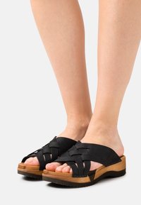 Svarta mockasin slidessandaler med vävda remmar, en plattform i träliknande material och en strukturerad gummisula. Tårna är synliga.