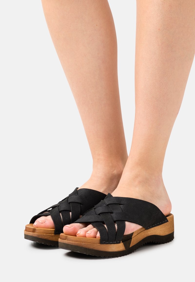 Svarta mockasin slidessandaler med vävda remmar, en plattform i träliknande material och en strukturerad gummisula. Tårna är synliga.