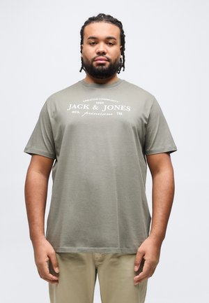 Mężczyzna z plecionymi włosami i brodą, ubrany w khaki spodnie i oliwkowo zieloną koszulkę marki Jack & Jones, stoi na tle jednolitego białego tła.