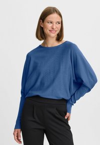 Pull en maille bleu surdimensionné avec un large col et des manches longues, associé à un pantalon noir ajusté avec une texture douce.