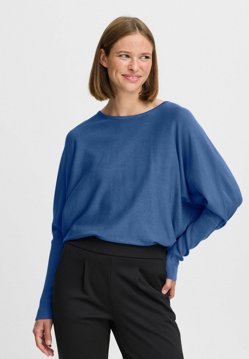 Pull en maille bleu surdimensionné avec un large col et des manches longues, associé à un pantalon noir ajusté avec une texture douce.