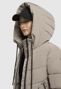 Beige Daunenjacke mit einer großen, gepolsterten Kapuze, schwarzem Reißverschluss, Kordelzügen und einer Akzenttasche. Getragen mit einer schwarzen Mütze.