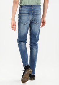 Personne portant un jean bleu délavé et des chaussures noires marchant de dos, sur un fond blanc.