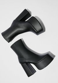 Par de botas de cuero negro con plataforma y tacones altos gruesos sobre fondo blanco.