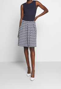 Granatowa bluzka bez rękawów o dopasowanej talii, w połączeniu z kremową spódnicą do kolan z zygzakowatym wzorem w granatowy i biały. Białe buty.