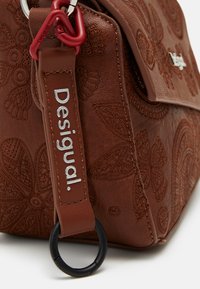 Borsa in pelle marrone con texture in rilievo floreale, dettagli in argento e un accento di hardware rosso. Presenta una tracolla robusta e un anello circolare.