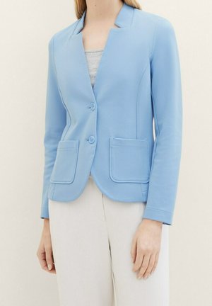 Femme portant un blazer bleu clair boutonné avec deux poches à l'avant, par-dessus un top gris et un pantalon blanc, sur un fond uni.