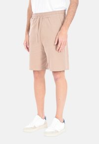 Pantaloni corti marrone chiaro, fino al ginocchio, con vita elastica e dettaglio con cordoncino. Realizzati in un tessuto morbido e testurizzato. Indossati con sneakers bianche.