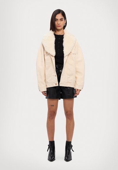 Chaqueta de mezclilla de gran tamaño color crema con un cuello de piel sintética, combinada con una blusa negra y pantalones cortos de cuero negro, llevada con botas negras hasta los tobillos.