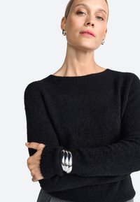 Schwarzer Wollpullover mit lockerer Passform, weitem Rundhalsausschnitt und langen Ärmeln; ausgestattet mit einem silbernen Armreif mit drei miteinander verbundenen Ringen.