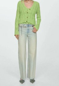 Zelený pletený svetr na knoflíky, kombinovaný se světle modrými džíny s širokými nohavicemi. Výrazné detaily zahrnují řetízkový pásek a ošuntělé lemy.