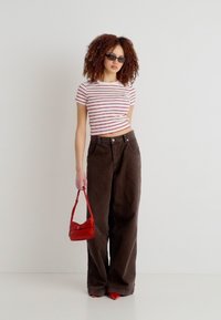 Γυναίκα με μπούκλες, φοράει μαύρα γυαλιά ηλίου, λευκό και κόκκινο ριγέ κοντό τοπ, φαρδιά καφέ παντελόνια, κόκκινα παπούτσια και κρατάει μια μικρή κόκκινη τσάντα.