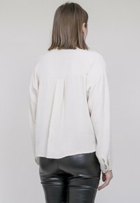 Blouse crème texturée à manches longues avec un plissé au dos. Associée à un pantalon en cuir noir brillant. Design simple, tons neutres.