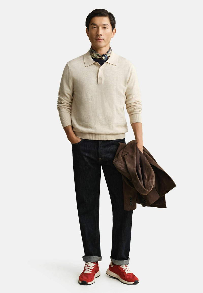 Homme portant un pull beige, un jean foncé, des baskets rouges, tenant une veste marron, debout devant un fond blanc.