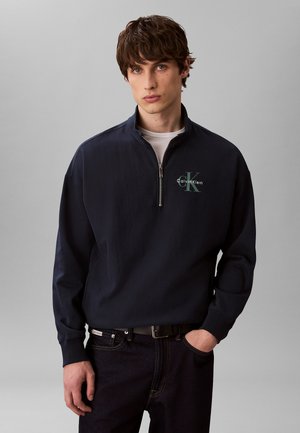 Młody mężczyzna w granatowej bluzie Calvin Klein z zamkiem do połowy, na białej koszulce i ciemnych jeansach, stoi na szarym tle.