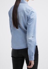 Light blue denim shirt with a button-up front, long sleeves, and a pointed collar. Features a slightly fitted shape and a smooth texture.