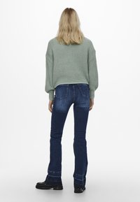 Lichtgroene gebreide trui met verlaagde schouders, gecombineerd met donkerblauwe flared jeans. Zwarte chunky laarzen en een gladde textuur zijn zichtbaar.