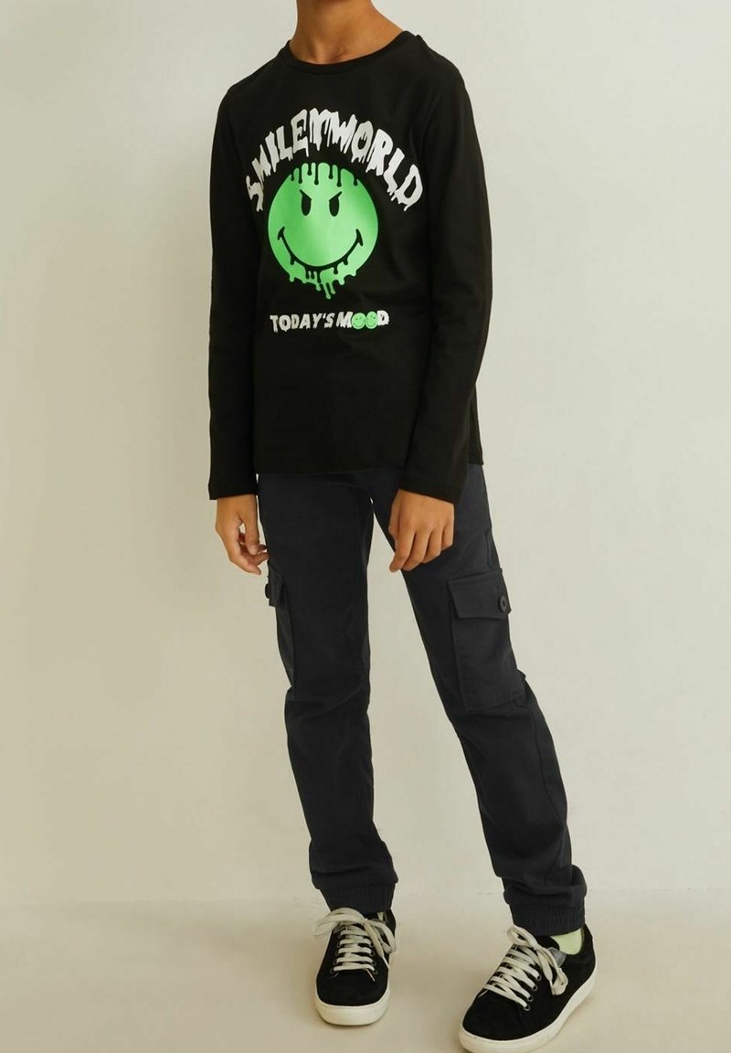 Enfant portant un t-shirt noir à manches longues avec un smiley vert dégoulinant, un pantalon cargo noir et des baskets noires à lacets blancs, debout contre un mur uni.