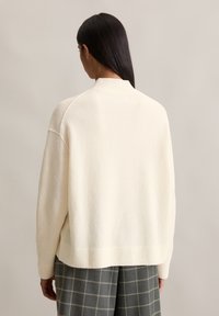 Crèmekleurige oversized trui met een geribde hals, zachte textuur, gestructureerde schouders en een afgeronde zoom, gecombineerd met een geruite grijze broek.