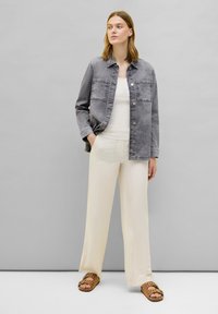 Giacca di jeans grigia con due tasche sul petto, abbinata a una canotta bianca e a dei pantaloni larghi crema, completata con sandali marroni.