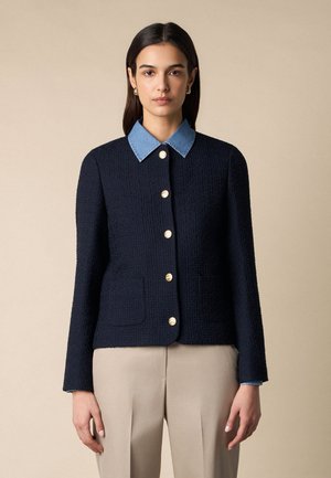 Žena v tmavě modré texturované bundě na knoflíky se zlatými knoflíky, pod kterou má modrou košili s límečkem, a béžových kalhotách na jednobarevném pozadí.