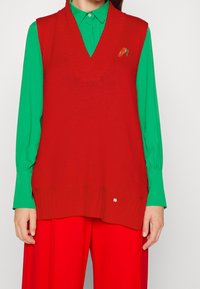 Veste rouge en tricot avec un décolleté en V, une texture côtelée et un détail de fraise brodé. Portée par-dessus une chemise verte à manches longues.