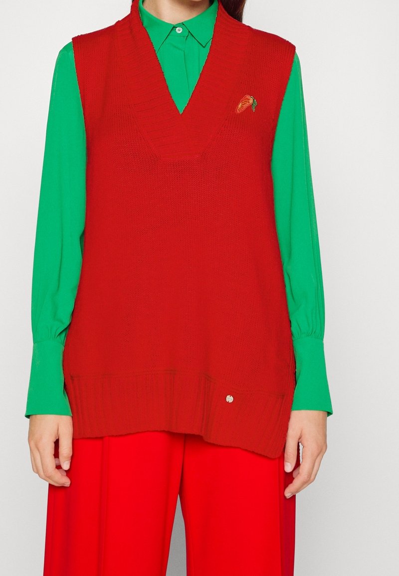 Veste rouge en tricot avec un décolleté en V, une texture côtelée et un détail de fraise brodé. Portée par-dessus une chemise verte à manches longues.