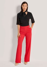 Zwarte blouse met een strikdetail, gecombineerd met felrode wijde broek en naaktsandalen, tegen een neutrale achtergrond.