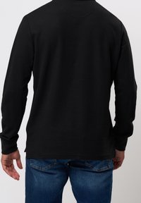 Uomo che indossa una maglietta nera a maniche lunghe e jeans blu, visto da dietro su uno sfondo chiaro e uniforme.