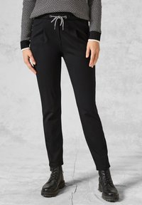 Svarta tapered byxor med elastisk midja och dragsko, med två sidfickor och en mjuk tygstruktur, kombinerade med svarta kängor.