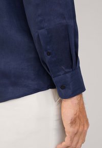 Camisa de lino azul marino con mangas largas, que presenta botones negros en los puños y una tela suave, ligeramente texturizada. Corte entallado en la cintura.