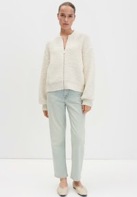 Cardigan a maglia strutturata color crema, con cerniera frontale, polsini e orlo a coste. Abbinato a jeans dritti blu chiaro e scarpe basse.