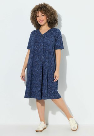 Frau steht an einer weißen Wand, trägt ein knielanges blau gemustertes Kleid und weiße Sneaker, lächelt mit lockigem Haar.