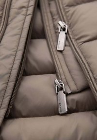 Gevoerde jas in een zachte taupe kleur met exposé ritsen en metalen hardware. Textuurstof met gestikte quiltpatroon.