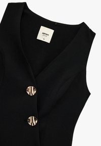 Chaleco negro sin mangas hecho de tela suave, con un escote en V profundo, dos botones redondos en tono dorado y una etiqueta que dice "Koton Business."