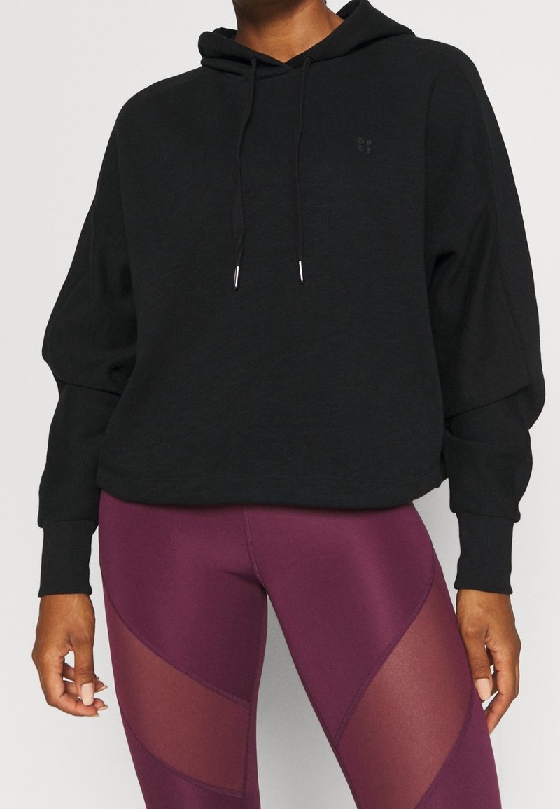 Personne portant un sweat à capuche noir avec cordons de serrage et un legging violet avec des empiècements transparents, vue des épaules aux cuisses sur un fond uni.