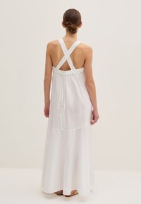 Abito maxi bianco con spalline incrociate, cintura elastica e gonna fluida. Texture liscia, design minimalista e dettaglio sul retro visibile.