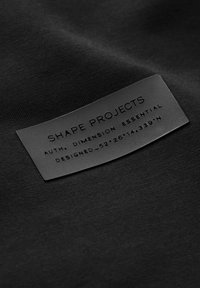 Sort stofmærke med præget tekst. "SHAPE PROJECTS" øverst, efterfulgt af "AUTH. DIMENSION ESSENTIAL" og koordinater nedenfor. Blød tekstur.