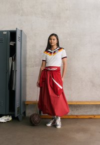 Λευκό κοντομάνικο πουκάμισο με πολύχρωμες διαγώνιες ρίγες, συνδυασμένο με μια κόκκινη μακριά φούστα. Ασημί αθλητικά παπούτσια και καφέ μπάλα μπάσκετ με δίχτυ.