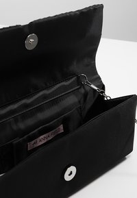 Černá látková peněženka s hladkou texturou, vybavená stříbrným zapínáním na patentku, vnitřními přihrádkami a štítkem přišitým uvnitř.