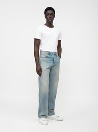 Jeune homme debout portant un t-shirt blanc à manches courtes, un jean bleu clair et des baskets blanches sur un fond blanc uni.